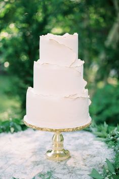 a white wedding cake sitting on top of a gold stand in front of some trees