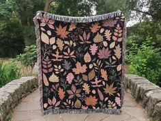 a black blanket with pink and green leaves on it sitting on a stone wall in front of some trees