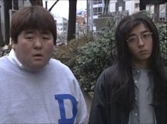 two young men standing next to each other in front of some bushes and trees on the sidewalk