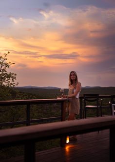 a woman standing on top of a wooden deck next to a table and chairs at sunset