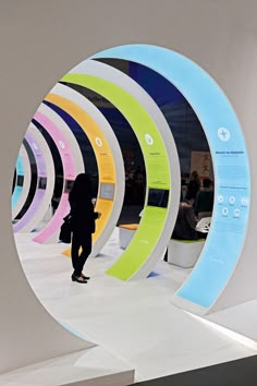 a woman is standing in front of a circular mirror with different colors on it and people are walking around the room