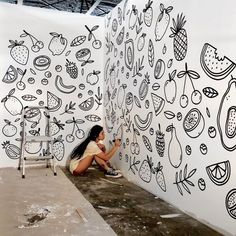 a woman sitting on the ground in front of a wall with fruit drawings painted on it