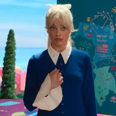 a woman with blonde hair wearing a blue sweater and white collared shirt standing in front of a map