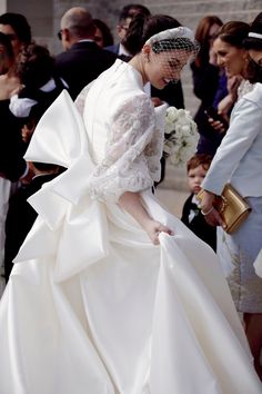 a woman in a white dress and veil walking down the street with other people behind her