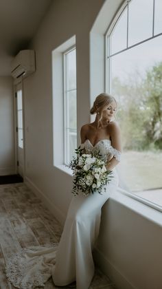 a woman in a wedding dress standing by a window with her bouquet on the windowsill