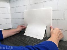 a person holding up a piece of paper on top of a wooden table next to a brick wall