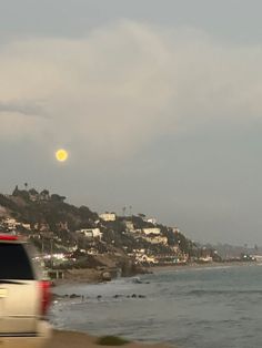 a car driving down the road next to the ocean with houses and hills in the background