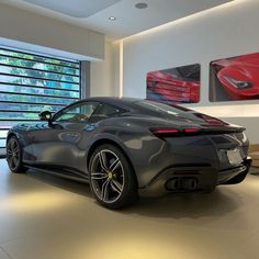 a grey sports car in a showroom with large windows and paintings on the wall