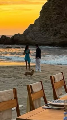 two people and a dog on the beach at sunset with an ocean in the background