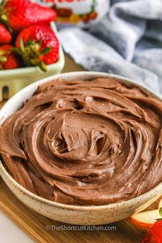 a bowl filled with chocolate frosting next to an apple and strawberries on a cutting board