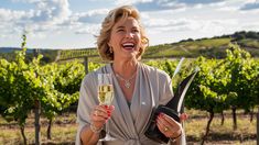 a woman holding a glass of wine in front of a bunch of vineyards and smiling at the camera