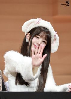 a girl with long brown hair wearing a white coat and holding her hand up in front of her face