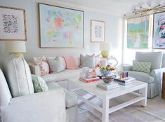 a living room filled with white furniture and paintings on the wall above it's coffee table