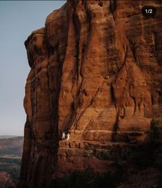 two people climbing up the side of a large rock formation with trees and mountains in the background