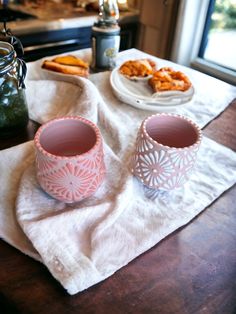 two pink cups sitting on top of a white towel next to a plate of food