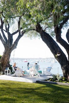 two people sitting on lawn chairs under large trees near the water with boats in the background