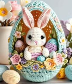 a crocheted bunny sitting in an egg with flowers and eggs on the side