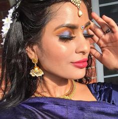 a woman with dark hair and blue eyeshadow wearing gold jewelry, holding her hand up to her face