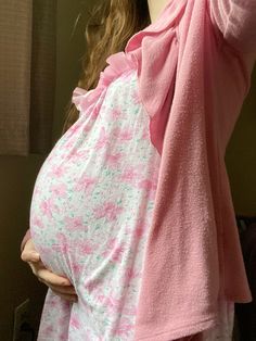 a pregnant woman wearing a pink robe and holding her hand up to her head while standing in front of a window