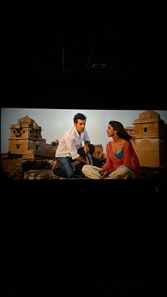 a man and woman sitting on the ground in front of a screen with an image of two people