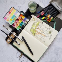 an open notebook sitting on top of a table filled with markers, pens and watercolors