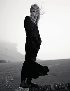 a woman standing on top of a stone wall next to a body of water with her hair blowing in the wind