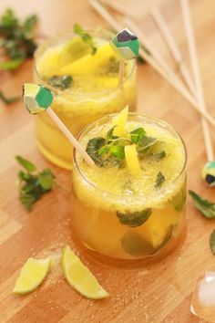 two glasses filled with drinks sitting on top of a wooden table next to lime wedges