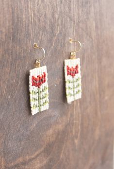 two small beaded earrings hanging from hooks on a wooden surface, one with red and green beads