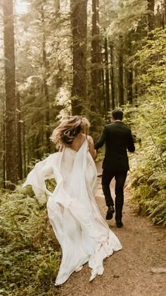 a bride and groom walking through the woods