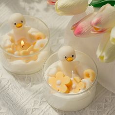 two small candles with ducks in them sitting on a white tablecloth next to flowers