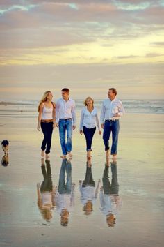 three people and a dog walking on the beach with their reflection in the wet sand