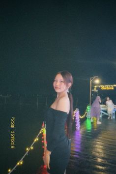 a woman standing on a pier at night with lights all around her and wearing a black off the shoulder dress