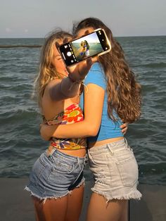 two young women are taking pictures with their cell phones by the water's edge