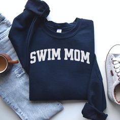 a sweatshirt with the word swim mom on it next to some shoes and a pair of jeans