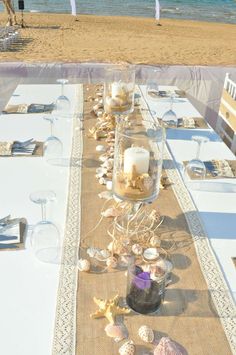 a table set up on the beach with shells and candles in glass vases next to it