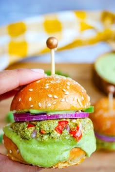 a hand is holding a small burger with green sauce and vegetables on it, while another hamburger sits in the background