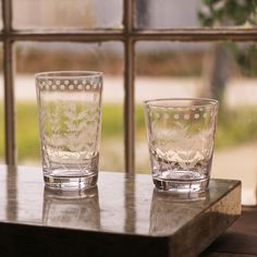 two glass cups sitting on top of a wooden table next to a window sill