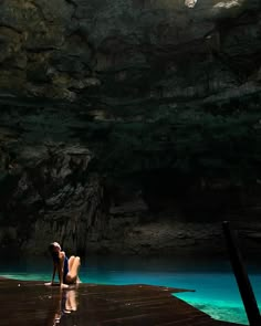 two people standing on a dock in front of a cave with blue water and cliffs