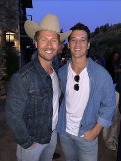 two men standing next to each other in front of a building at night, one wearing a cowboy hat and the other smiling