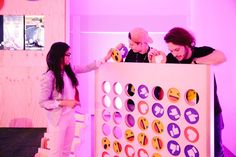 two people standing next to a giant board game in a room with purple lighting and pink walls