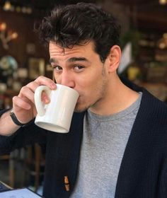 a man sitting at a table drinking from a coffee cup with his hand on the mug