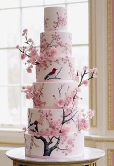 a three tiered cake with pink flowers and birds on the top is sitting on a table in front of a window