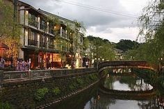 people are walking on the bridge over the water near buildings and trees in the evening