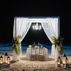 a table set up on the beach at night for a formal dinner with candles and flowers