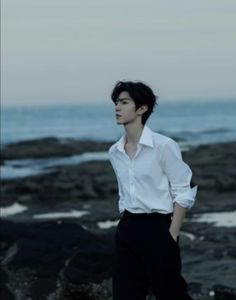 a young man standing on top of a rocky beach next to the ocean wearing a white shirt and black pants