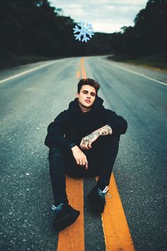 a young man sitting on the side of an empty road with his hands in his pockets