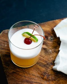 a small glass filled with liquid and a cherry garnish sitting on top of a wooden cutting board