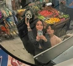 two women taking a selfie in front of a fruit stand with a skeleton mask on