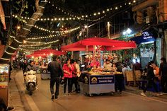 people are walking down the street at night with lights strung overhead and food stalls on both sides