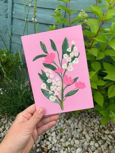 a hand holding up a pink card with flowers on it in front of some plants